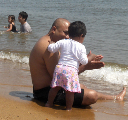 3 photos show an Arab man sitting in the shallow water, and deeper in the water is a Chinese man squatting.  Each man is with a little girl about 2-3 years old.  The Arab girl first stands behind her father, then sits on his lap.  The Chinese father is holding the girl and pulling her through the water.